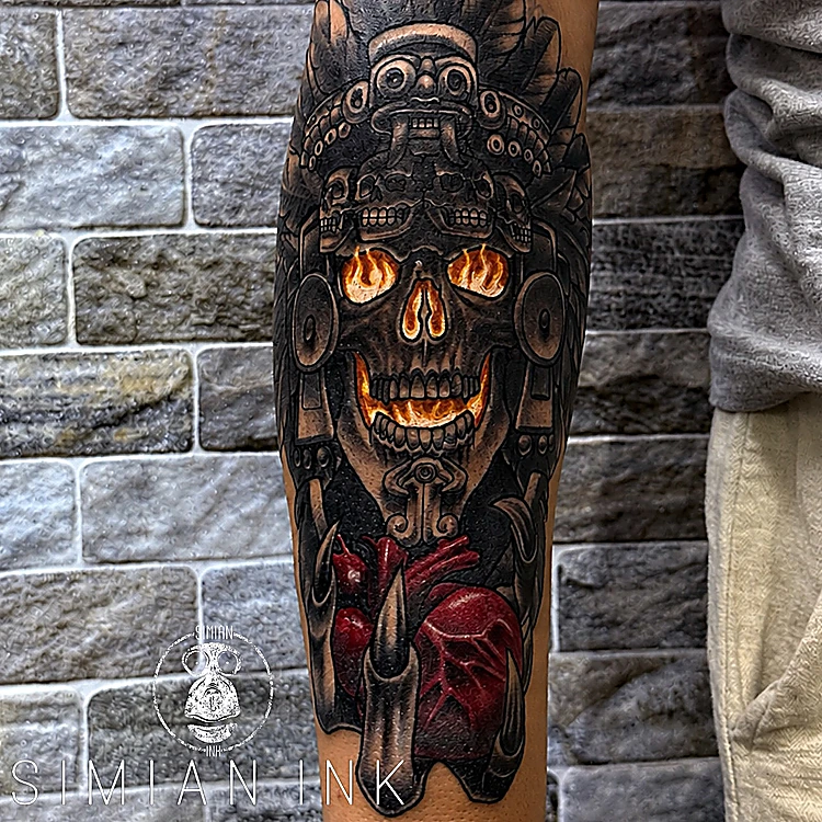 Tatuaje de calavera prehispánica con ojos de fuego sobre un corazón rojo, blackwork con color en el antebrazo