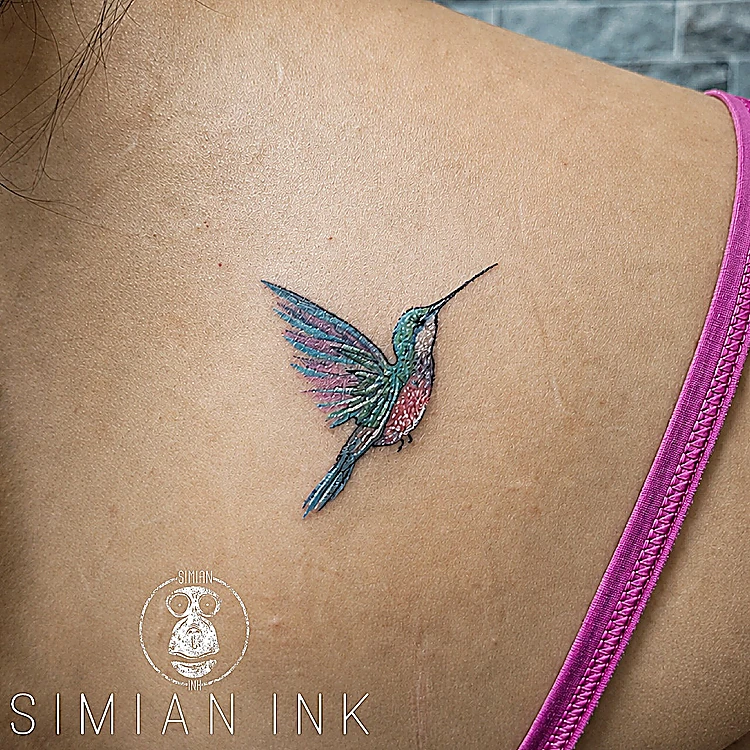Tatuaje pequeño de colibrí a color en azul, verde y magenta sobre el omóplato