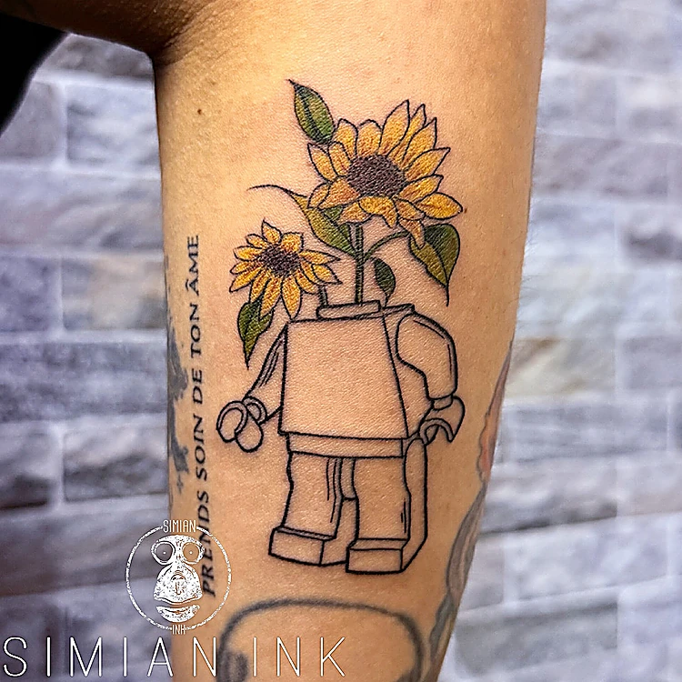 Tatuaje de una figura de Lego con girasoles en lugar de cabeza, en línea y color suave sobre el brazo